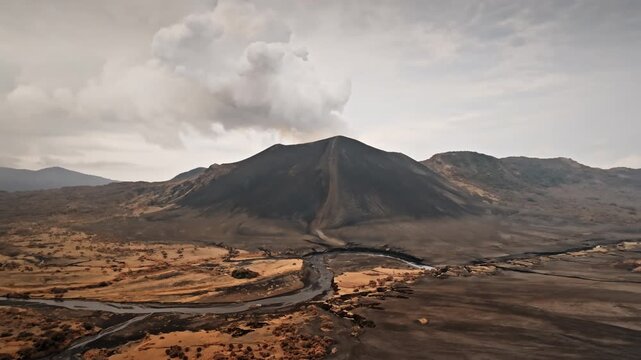 Mount Yasur volcano on Tanna Island is actively emitting smoke and ash into the cloudy sky, creating rugged volcanic landscape with winding river and dark black sand beach below. Aerial view panorama