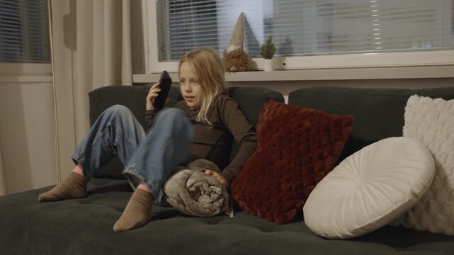 Young blonde girl sitting on a comfortable sofa at home, talking on a smartphone while holding a grey blanket, surrounded by pillows with window blinds behind her
