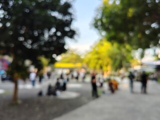 Blurred public park background with people relaxing under green trees during sunny daytime outdoor event © Ricko Destu