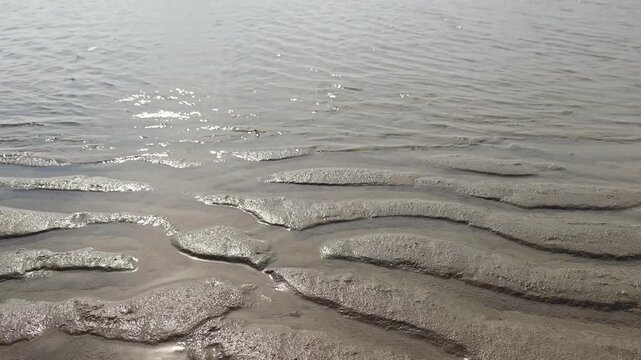 A wide-angle landscape shot of natural wavy sand ripples on a coastal mudflat beach during low tide, reflecting bright sunlight against a soft cloudy sky at the horizon.