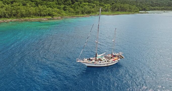 Sailboat resting on calm, deep blue and turquoise tropical waters near lush green coastline. Adventure, travel, and tranquility in the South Pacific, Tegua island, Vanuatu. Aerial view drone panorama