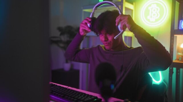Man puts on headset while hands hover over keyboard and smiles at monitor in gaming studio with neon signs and microphone visible; focused fun.