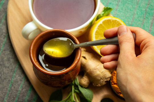 Hand holding spoon with honey over clay jar next to cup of herbal tea lemon and ginger root.