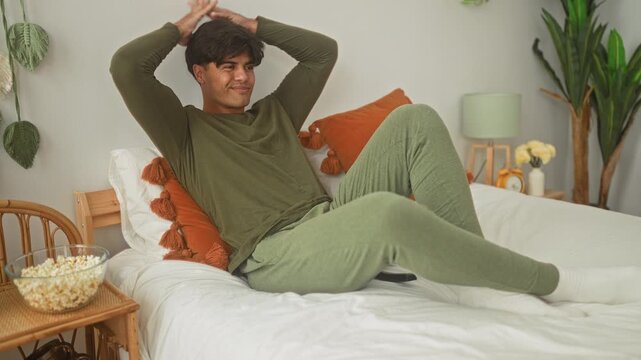 Man on bed with bowl of popcorn and remote, hands behind head in green loungewear, reclining on pillows in studio; relaxed comfort.