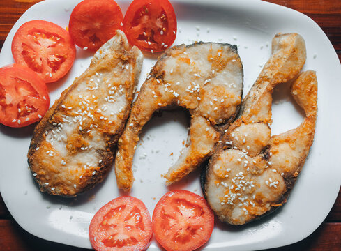 Pieces of sesame-fried fish served on a white plate with sliced tomato. A perfect dish for breakfast, lunch, or dinner.