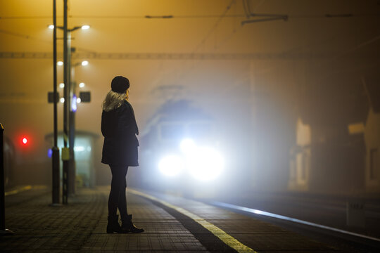 Woman looking at arriving train at empty railway station at night. City life and commute by public transportation