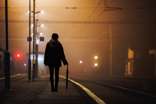 Railway station at night. Dark silhouette of woman with umbrella waiting for train on empty railroad station platform