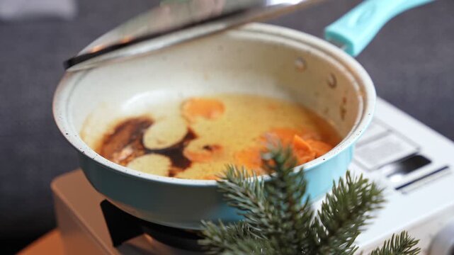 a hand lifting a glass lid off a blue frying pan on a portable stove. The indoor cooking scene features a small pine plant and ring light reflections on the cookware.