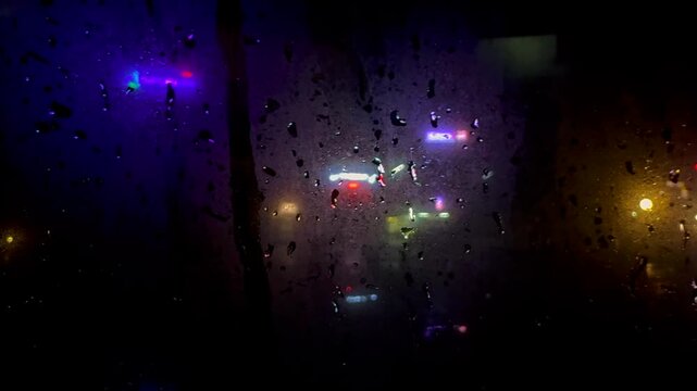 Abstract nighttime city lights viewed through the rain covered window of a moving vehicle, with water droplets, motion blur and colorful reflections creating a moody urban atmosphere
