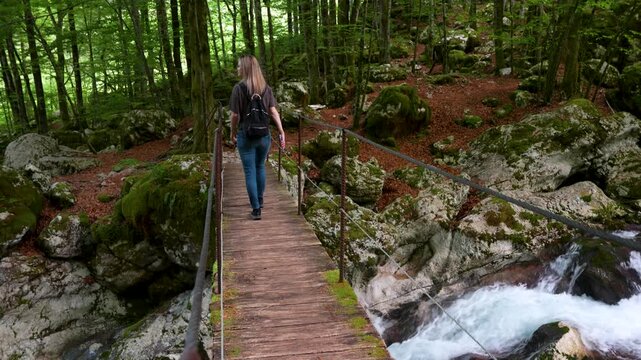 A young woman with a backpack walks steadily across a rustic wooden bridge over a rushing mountain stream in the lush &Scaron;unik Water Grove forest of Lepena Valley, Slovenia.