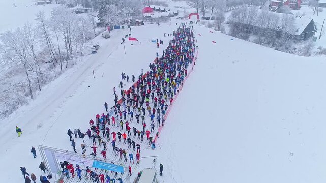 Aerial view of a large group of cross-country skiers gathered at the starting line of a winter race on a snow-covered field in Latvia.
