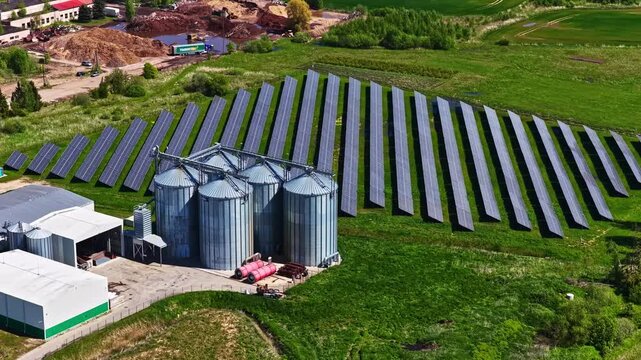 Aerial view of solar farm with rows of photovoltaic panels next to large metal grain silos and industrial buildings in the green countryside of Riga, Latvia.