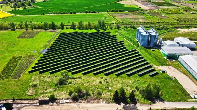 Aerial view of solar farm with rows of photovoltaic panels situated in a green field next to agricultural buildings and silos in Riga, Latvia.