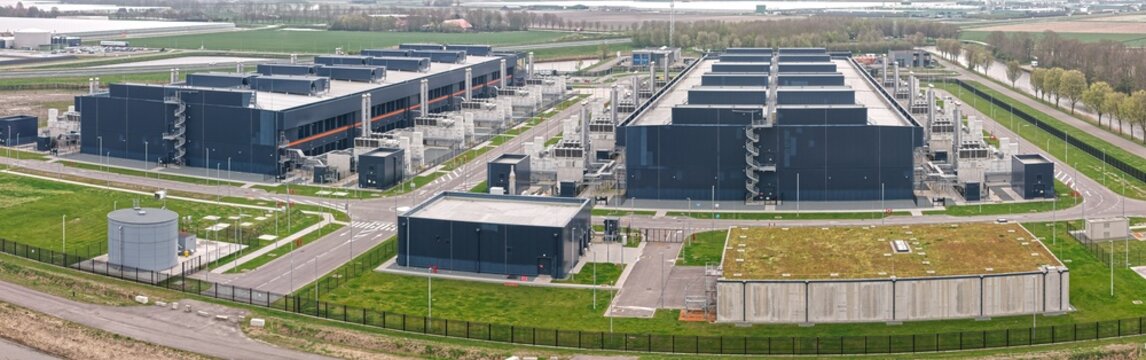Aerial view of a large modern data center campus with dark industrial buildings, rows of back-up energy generators, and green surroundings in Middenmeer, Noord-Holland, Netherlands.