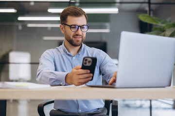 Obraz premium Young businessman multitasking, checking smartphone and working on laptop in modern office, managing digital tasks