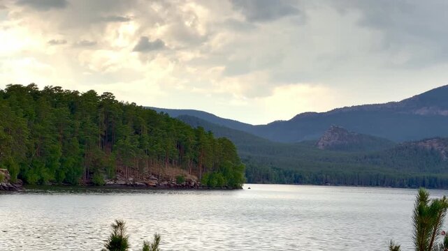Scenic mountain lake bordered by a dense pine forest in Burabay National Park, Kazakhstan. Peaceful natural landscape with calm water, rolling hills and evergreen trees under a dramatic cloudy sky