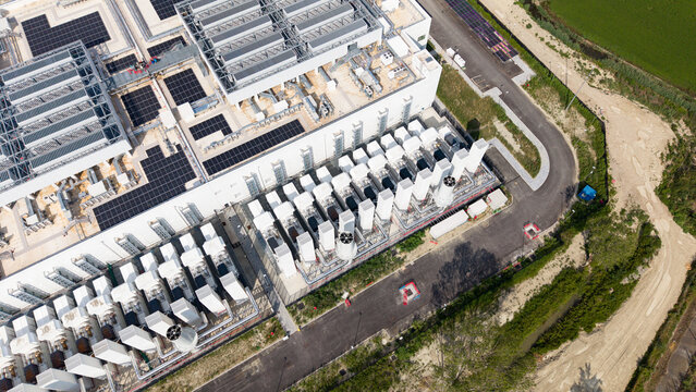 Aerial view of a modern data center campus featuring solar panels on the roof and industrial cooling units, situated near a paved road in Melegnano, Lombardy, Italy.
