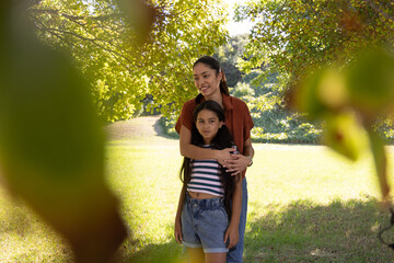 Obraz premium African American mother embracing youth daughter in park under large tree, wearing rust shirt