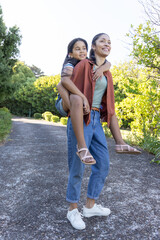 Obraz premium African American mother carrying teenage daughter on back walking along paved park path past bushes