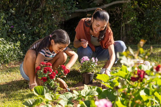African American mom and adolescent daughter planting red mums in backyard using trowel and gloves