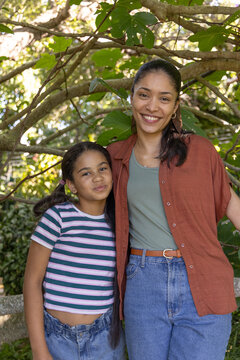 African American mother and youth daughter smiling under leafy canopy in garden, wearing belt