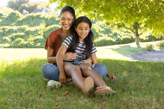 African American mother and daughter sitting on grass under tree, holding child with denim overalls