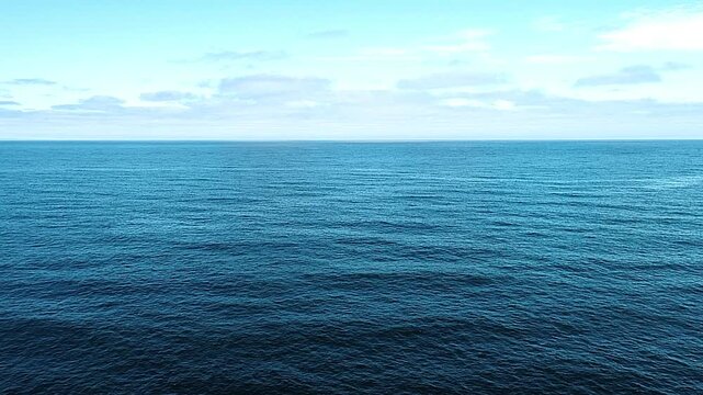 Ocean surface extends toward the horizon under a bright sky with soft scattered clouds.