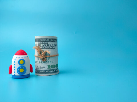 A toy rocket stands next to a rolled US hundred-dollar bill tied with twine on a blue background