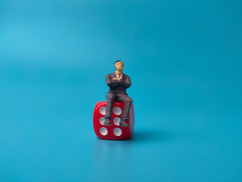 A miniature businessman figurine sits calmly on top of a red die against a solid blue background