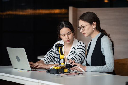 Robotics Education with a female teacher and Asian student using a laptop beside a yellow robotic arm and electronic equipment in a STEM classroom