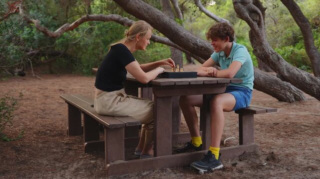 A mother and her fifteen year old teenage son play chess together at a wooden picnic table in a pine forest park, smiling and moving chess pieces on a sunny day