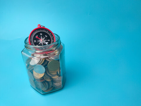 A compass sits at the top opening of a clear glass jar filled with mixed coins on blue