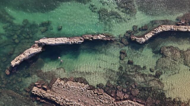 Aerial view of the rocky coastline and clear turquoise waters at Praia das Avencas with swimmers in the sea in Cascais, Lisboa, Portugal.