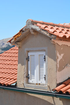 A close up view shows a small dormer window with white closed shutters on a weathered building roof with terracotta tiles.