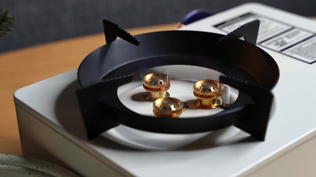 a white portable gas stove with a black pan support sitting on a wooden table indoors. Bright blue flames burn from the gold brass burners during daytime meal preparation.