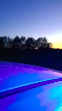 Red and blue police lights flashing and reflecting on the wet rooftop of a car at a crime scene, with a beautiful blurry sunset and dark trees creating a dramatic bokeh effect in the background