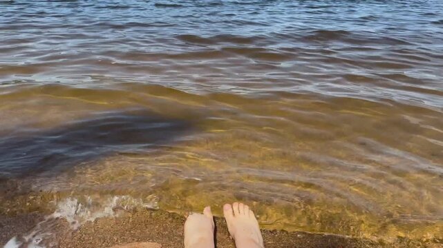 Bare feet touching the clear water at the edge of a sandy lake shore on a warm summer day. Relaxing outdoor moment, vacation atmosphere, freshwater landscape and peaceful nature experience.