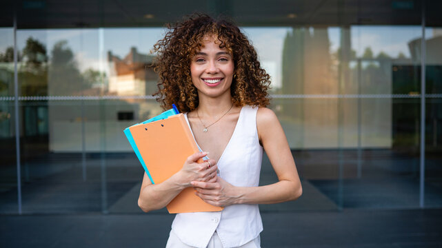 Confident young businesswoman holding folders outside office building