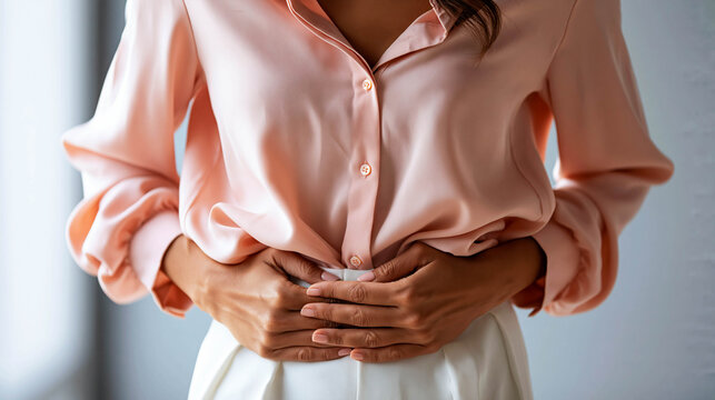 A young woman in a pink blouse holds her stomach and writhes in pain on a light background, representing the concept of abdominal pain, digestive disorders, and menstrual cramps.