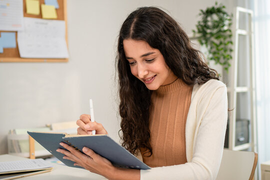 Young Hispanic woman writing while sitting at table in living room at home. 