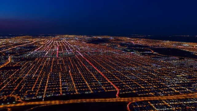 Aerial night city grid with illuminated streets, highways, traffic light trails, coastal horizon, deep blue sky, amber glow, dense urban blocks, and modern metropolitan scale