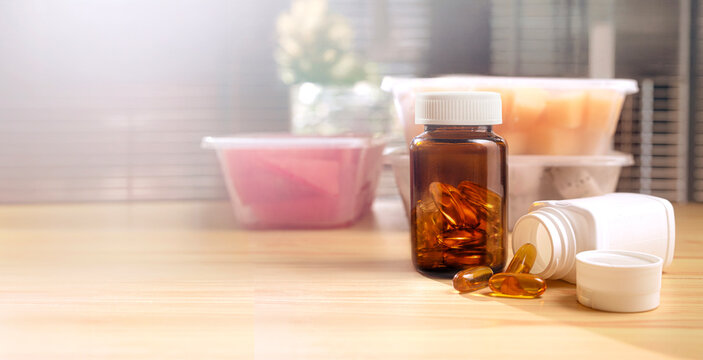 Amber vitamin supplement bottles with softgel capsules placed beside healthy meal prep containers on a wooden table. Healthy nutrition, wellness, dietary supplements