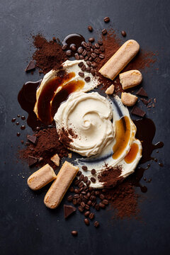 A beautiful flat lay of classic tiramisu ingredients with swirls of mascarpone cream, dark espresso coffee, cocoa powder, ladyfinger biscuits, and chocolate on a dark slate background.