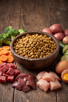 A dark wooden bowl filled with dry dog kibble surrounded by fresh raw ingredients including chicken, beef, liver, raw egg yolk, carrots, broccoli, potatoes, and zucchini on a rustic table.