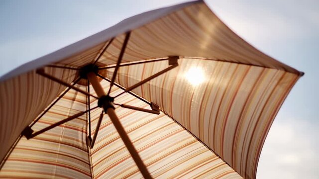Low-angle close-up of striped beach umbrella underside with sun glare through translucent canopy, showing central pole, ribs, connectors, and warm summer backlight over outdoor leisure scene