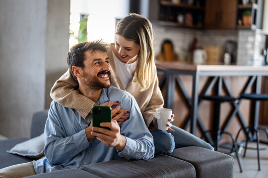 Happy couple enjoying morning coffee at home while browsing their smartphone, showing love