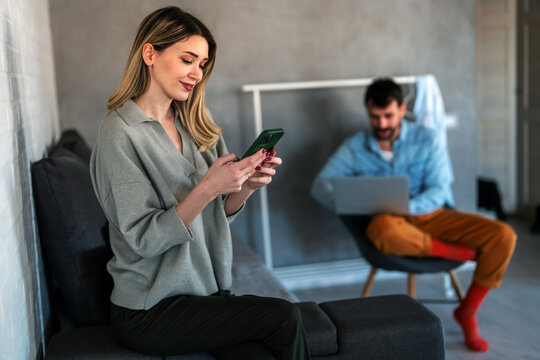 Smiling caucasian woman using her smartphone while her partner works in the background, home office