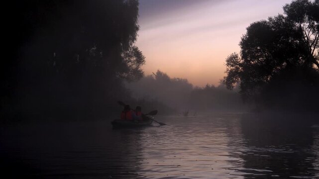 Couple paddling kayak on serene river during beautiful misty morning, surrounded by silhouettes of trees and gentle glow of rising sun creating tranquil atmosphere. Romantic activity of a couple