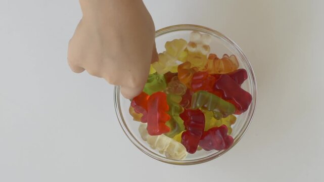 Child hand choosing gummy bear from glass bowl filled with colorful jelly candies on white background. Kid carefully selects favourite colour before taking sweet. Eating choices, decision making 
