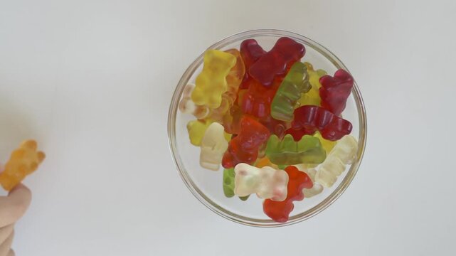 Child hand takes orange gummy bear from colourful candy bowl placed on white background. Bright overhead composition focuses on colour recognition, playful learning and favourite sweet selection